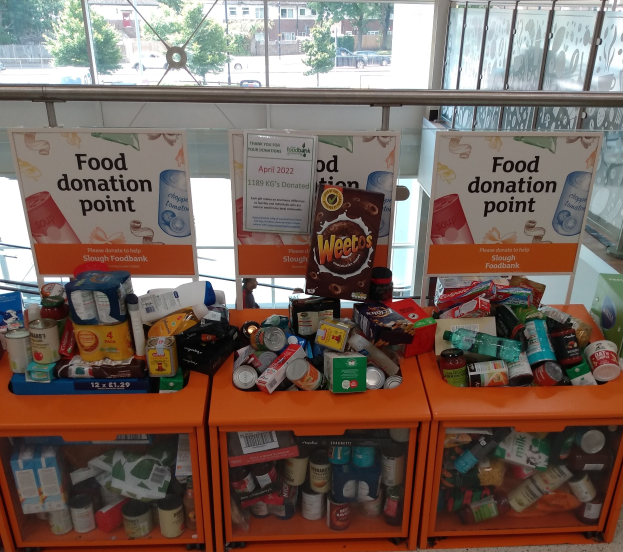 Orange Schränke mit Lebensmittelspenden gefüllt, darunter Flaschen, Dosen und Kartons, mit Informationsschildern im Hintergrund; Glasfenster zeigen Fahrzeuge, Bäume, Gebäude und Himmel.