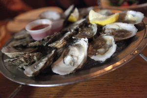 Ein Teller mit Muscheln, die nebeneinander angeordnet sind, ein Becher mit Sauce in der Mitte und zwei Zitronenstücke daneben, mit zusätzlichen Tellern im Hintergrund auf dem Tisch.