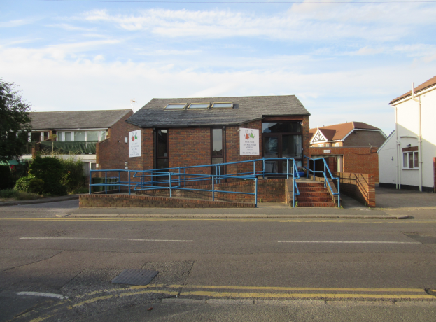 Ein Schulgebäude mit einer blauen Reling davor, umgeben von Häusern mit Fenstern und Türen, Treppen mit Relings, Schildern mit Text, Pflanzen, Bäumen und einem bewölkten Himmel im Hintergrund.