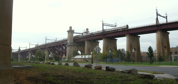 Eine Bahn überquert eine Brücke mit Säulen, Strommasten mit Drähten, Schilder, einen Zaun, Gras, Steine, eine Straße, Bäume, Gebäude und einen bewölkten Himmel im Hintergrund.