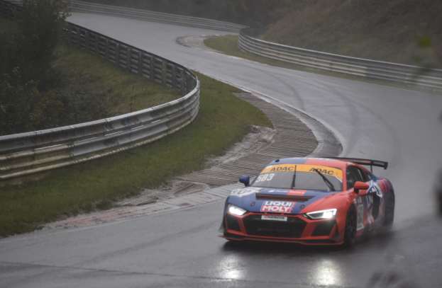 Audi R8 LMS GT3 Rennauto auf der Nürburgring 24 Stunden Strecke, umgeben von grünem Gras und Bäumen mit einem Geländer im Hintergrund.