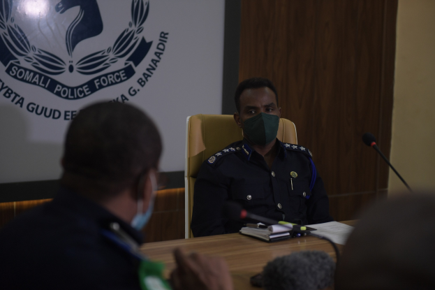 Zwei Polizeibeamte in Masken sitzen an einem Tisch mit Mikrofonen, Büchern und anderen Gegenständen und schauen auf einen Wandbildschirm mit Text und einem Emblem.