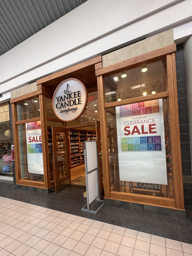 Storefront with a "Yankee Candle Clearance Sale" sign, glass walls and doors, and illuminated shelves inside displaying various items.