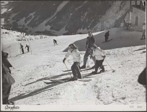 Gruppe von Menschen, die eine schneebedeckte Piste mit Ski-Stöcken hinunterfahren, mit Hügeln und einem Gebäude im Hintergrund und Text unten.