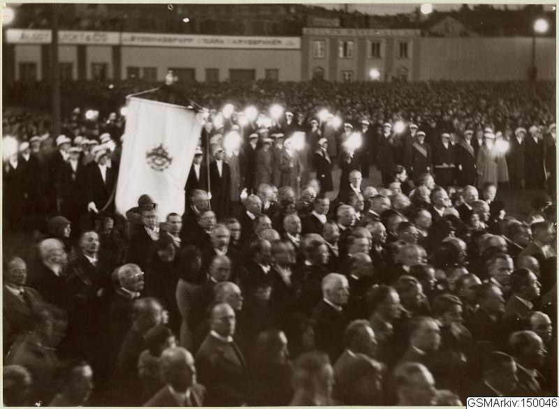 Schwarzes und weißes Foto einer großen Menge vor einem Gebäude, mit einer Person, die eine Fahne hält, und Text, der unten 'Gustav Klimts Rede an der Berliner Mauer' lautet.