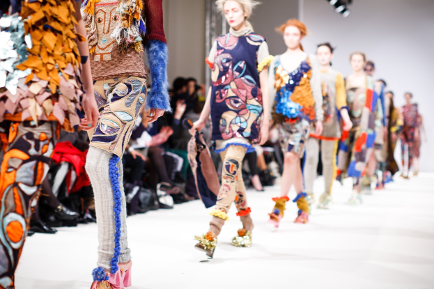Eine Gruppe Von Frauen Geht Den Laufsteg Bei Der London Fashion Week entlang, Mit Sitzenden Zuschauern Und Bühnenbeleuchtung Im Hintergrund.