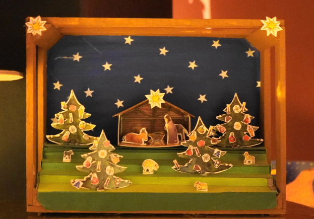 Ein hölzerner Krippenkasten auf einem Tisch, mit einer Lampe links und einer Wand im Hintergrund, dekoriert mit Weihnachtsbäumen, Tieren und einem Haus.