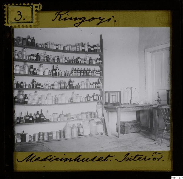 Schwarz-weiß-Foto einer alten Apotheke mit Regalen voller Flaschen, einem Tisch mit Gegenständen, einem Stuhl, einer Tür und einer Wand, mit Text unten.
