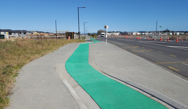 Eine Straße mit einem grünen Fahrradweg an der Seite, Gras auf der linken Seite, Verkehrskegel auf der rechten Seite, Laternenpfähle, Bäume, Gebäude und einen klaren blauen Himmel im Hintergrund.