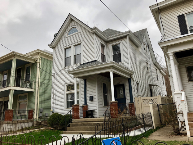 Reihe von Häusern mit Fenstern, Türen, Säulen, Geländern, Treppen und Dächern, umgeben von einem Metallzaun, Gras, Pflanzen, Bäumen und einem bewölkten Himmel, mit einem Mietschild am Boden, das die Adresse 5901 S Elmwood Ave, Chicago 60618 angibt.