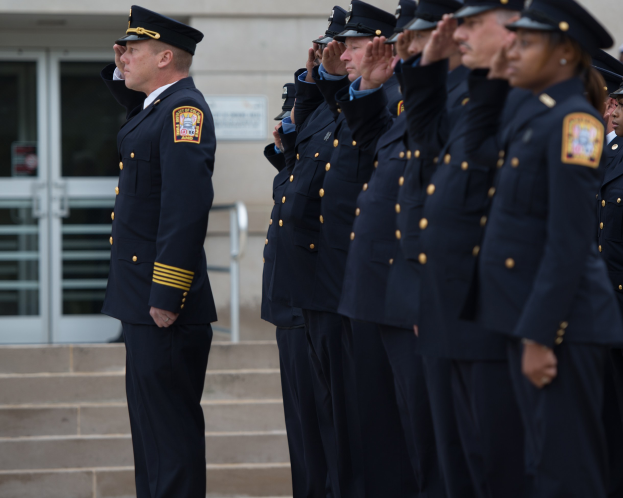 Gruppe von Polizisten in Uniform und Mützen, die in Formation vor einem Gebäude mit Glastüren und Treppen stehen und salutieren.