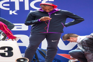 Eine Person, die strahlt und ein Mobiltelefon hält, mit einer weiteren Person rechts daneben, einem Rucksack, einem Megafon unten, einer Wand im Hintergrund und einer weiteren Person links.