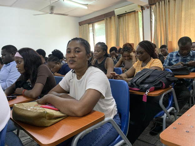Eine Gruppe von Studenten sitzt an Tischen in einem classroom an der Universität von Ghana, mit Taschen und Gegenständen auf den Tischen, Fenstern mit Vorhängen, einer Klimaanlage und Deckenventilatoren und -leuchten im Hintergrund.