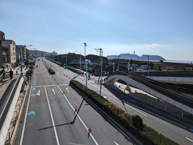 Straße mit Verkehrskegeln, eine Brücke mit Geländern, Laternenpfähle, Gebäude mit Fenstern und einige Menschen; Hügel und bewölkter Himmel im Hintergrund, Teil des Takayama-Brücken-Projekts.