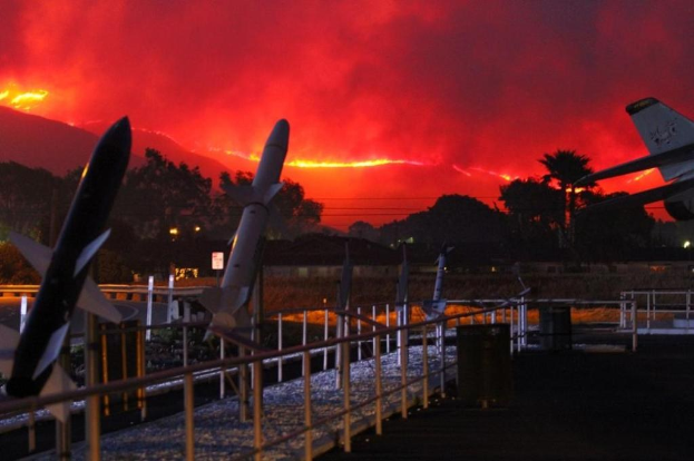 Ein Flugzeug fliegt über eine Start- und Landebahn mit Raketen, umgeben von Geländern, Mülltonnen, Bäumen, Laternenmasten und Gebäuden, mit Rauch von den Kalifornischen Waldbränden im Hintergrund.