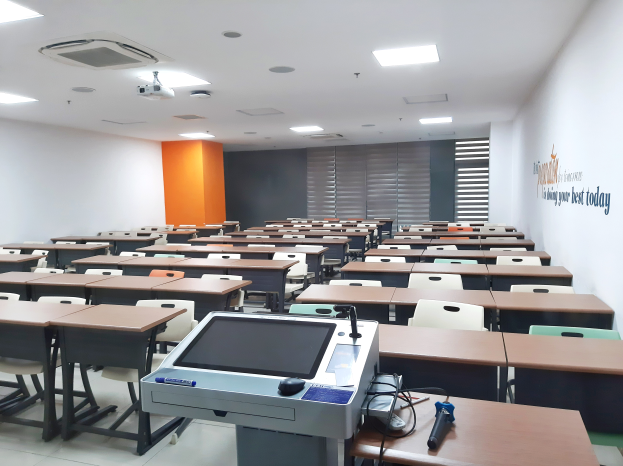 Ein Klassenzimmer mit Tischen und Stühlen in Reihen, einen deckenmontierten Projektor und verschiedene Gegenstände auf den Tischen sowie Text an der rechten Wand, der darauf hinweist, dass es sich um eine Privatschule in Singapur handelt.
