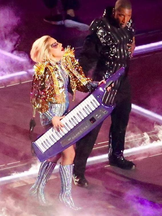 Lady Gaga und Kanye West bei der Halbzeitshow des Super Bowl auf der Bühne, mit Lady Gaga in einem farbenfrohen Kleid und einem Instrument spielend und Kanye West in einem schwarzen Outfit neben ihr, unter hellen Bühnenlichtern mit Rauch im Hintergrund.