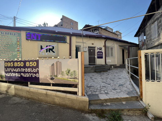 Gebäude mit einem Metallzaun und Tor, ein Schild mit der Aufschrift 'hr Immobilienbüro zur Miete in Phnom Penh', ein Weg, Pfosten, Drähte, Pflanzen, eine Baumgruppe und ein bewölkter Himmel.