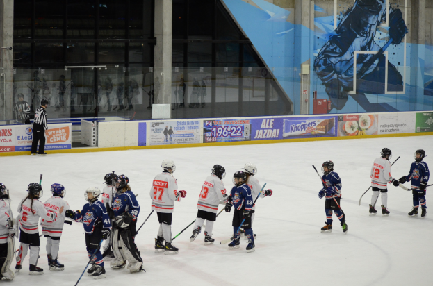 Junge Menschen in Helmen und Hockey-Ausrüstung spielen Eis Hockey auf einer Indoor-Eisfläche mit Bannern, Säulen und Glaswänden im Hintergrund.