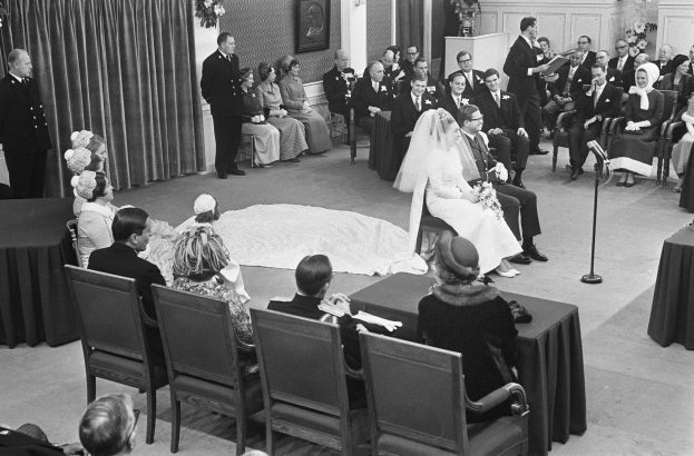 Schwarzes und weißes Foto einer Hochzeitszeremonie mit sitzenden und stehenden Menschen, einem Mann mit Mikrofon, einer Frau mit einem Strauß Blumen, einem Podium mit Mikrofon, Blumenschmuck, einem Vorhang, einem gerahmten Foto an der Wand und einer Tür im Hintergrund.