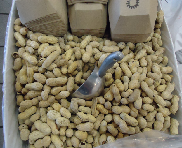 Eine Schachtel Erdnüsse auf einem Tisch mit einer Plastikabdeckung und einem Löffel daneben, umgeben von zusätzlichen verstreuten Erdnuss-Schachteln.