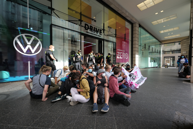 Menschen sitzen vor einer Volkswagen-Niederlassung auf dem Boden und halten ein Transparent mit Text während einer Demonstration, mit Glaswänden und Logo auf dem Gebäude.
