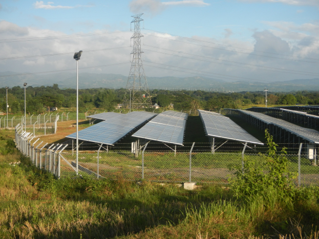 Luftaufnahme eines Solarpanelfelds, das von einem Zaun, Gras, Pflanzen und Bäumen umgeben ist, mit einem Übertragungsturm und Stromleitungen im Hintergrund bei bewölktem Himmel.
