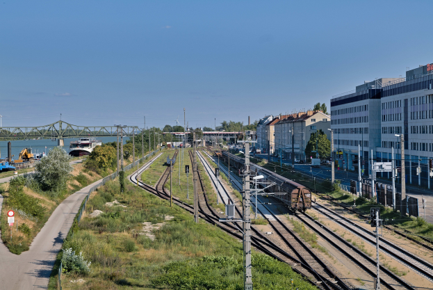 Ein Zug fährt auf Schienen neben einer Stadtlandschaft mit Infrastruktur und Grünfläche unter einem bewölkten Himmel.