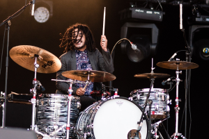Ein Mann mit Dreadlocks, der auf der Bühne Schlagzeug spielt und Drumsticks mit Mikrofonstativen vor sich hält.