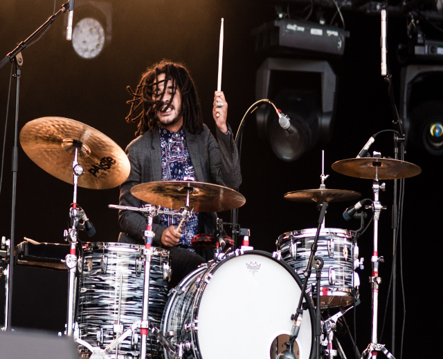 Ein Mann mit Dreadlocks, der auf der Bühne Schlagzeug spielt und Drumsticks mit Mikrofonstativen vor sich hält.