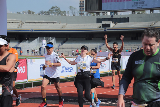 Gruppe von Menschen, die auf einer Stadionbahn laufen, mit Bannern, einem Display, einem Gebäude, Bäumen und einem bewölkten Himmel; einige Läufer tragen Turnschuhe und Hüte.
