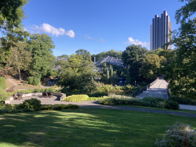 Ein Park mit grünem Gras, Bäumen, Pflanzen, Blumen, Bänken und einem Weg, vor Buildings und einem bewölkten Himmel.