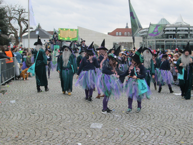 Eine Gruppe von Menschen in Hexenkostümen, die schwarze und lila Kleider und Hüte tragen und die eine Gasse entlanggehen, während einige Fahnen halten, neben einem Geländer mit Gebäuden, Bäumen und einem klaren blauen Himmel im Hintergrund.
