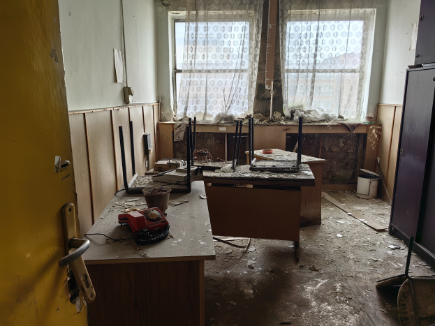 Leerer Raum mit einer Tür auf der linken Seite, einem Schrank auf der rechten Seite und einem Fenster mit Vorhängen im Hintergrund; ein Tisch in der Mitte hält verstreute Gegenstände und suggeriert ein verlassenen Büro.