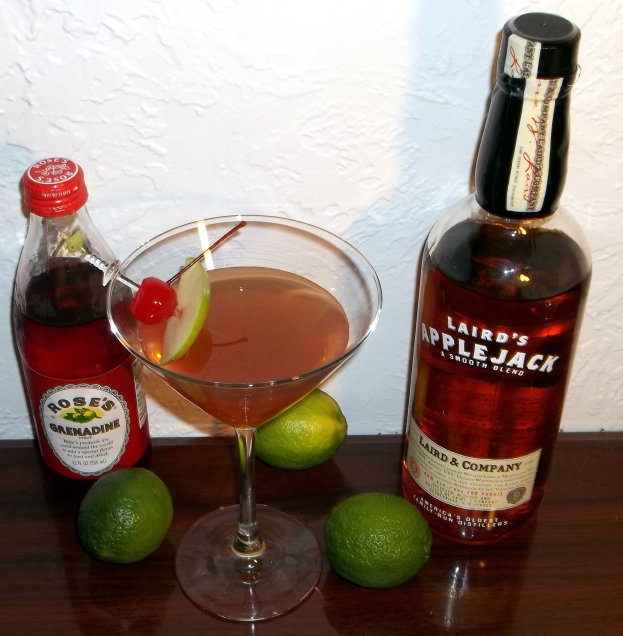 Martini in a coupe glass with cherry and two lime garnishes on a table next to a liquor bottle, against a wall background.