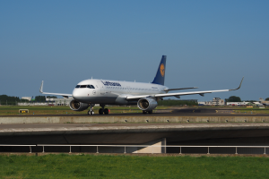 Ein Lufthansa Airbus A330-300 auf der Rollbahn am Frankfurter Flughafen, umgeben von Gras und einem Geländer, mit Gebäuden, Bäumen und einem klaren blauen Himmel im Hintergrund.
