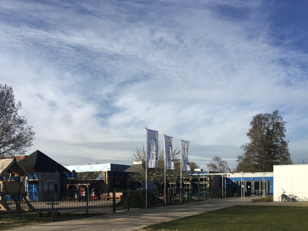 Großes Gebäude mit einem grasbewachsenen Bereich davor, umgeben von einem Metallzaun, mit einem Weg, Bäumen, Pfosten, Bannern, einem geparkten Fahrrad, nahegelegenen Häusern und einem bewölkten Himmel, als neuer Spielbereich für Kinder an der Schule identifiziert.