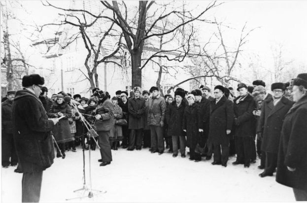 Eine Gruppe von Menschen in Mänteln und Mützen, einige mit Musikinstrumenten, steht im Schnee vor einer Menge, mit Bäumen und Gebäuden im Hintergrund, dargestellt in Schwarz-Weiß.