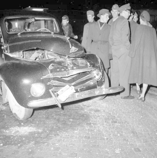 Schwarz-weiß-Foto eines schwer beschädigten Autos am Straßenrand mit eingedellter Motorhaube und zersplitterten Fenstern, umgeben von besorgten Menschen, die es inspizieren.