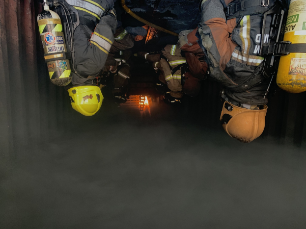 Feuerwehrleute in Schutzanzügen und Atemschutzgerät arbeiten in einem rauchgefüllten Tunnel, mit einer Lichtquelle im Hintergrund.