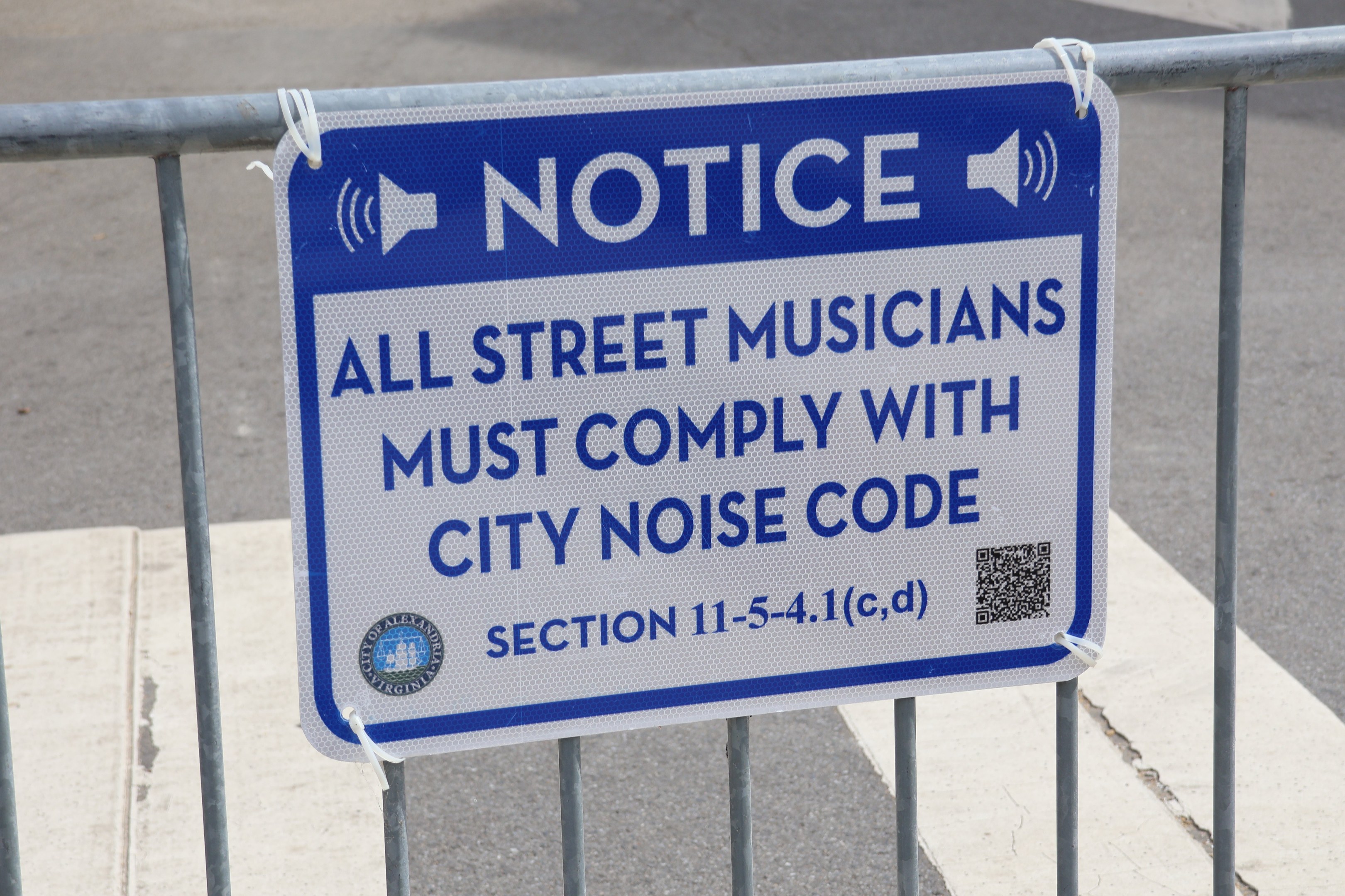 Anschlagtafel an einem Zaun mit einem Schild, auf dem "Anschlag: Alle Straßenmusiker müssen die Stadtlärmordnung einhalten" steht und eine Straße im Hintergrund.