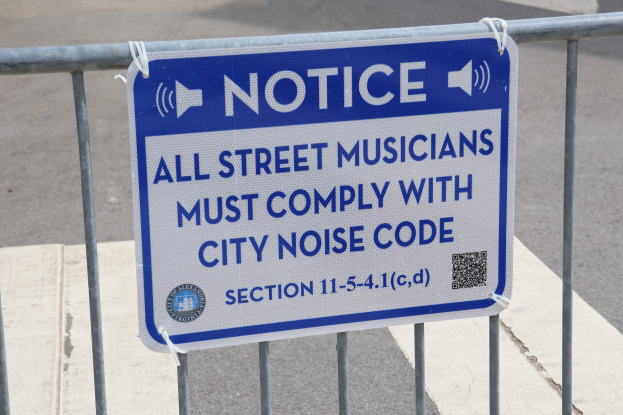 Anschlagtafel an einem Zaun mit einem Schild, auf dem "Anschlag: Alle Straßenmusiker müssen die Stadtlärmordnung einhalten" steht und eine Straße im Hintergrund.