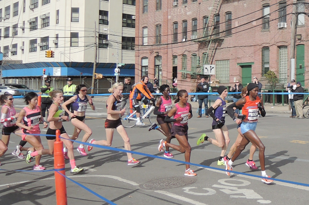 Gruppe von Menschen, die bei einem Marathon auf einer Straße mit Gebäuden, Strommasten, Ampeln, Schildern, Fußgängern, einem Radfahrer, Bäumen und einem klaren blauen Himmel laufen.