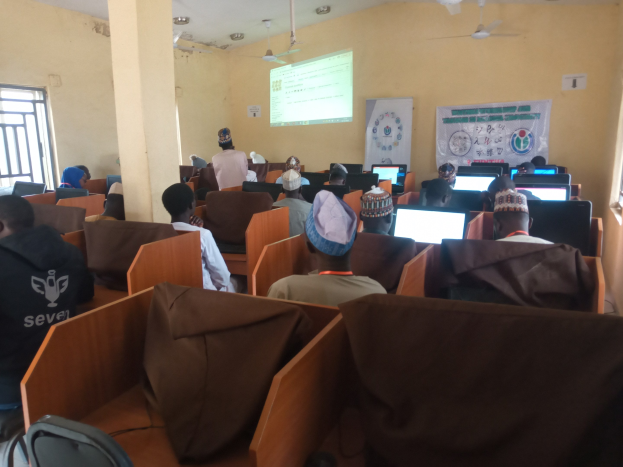 Eine Gruppe von Menschen sitzt an Computern in einem Seminarraum mit Deckenventilatoren, einem Bildschirm, einer Wand und Fenstern auf beiden Seiten.