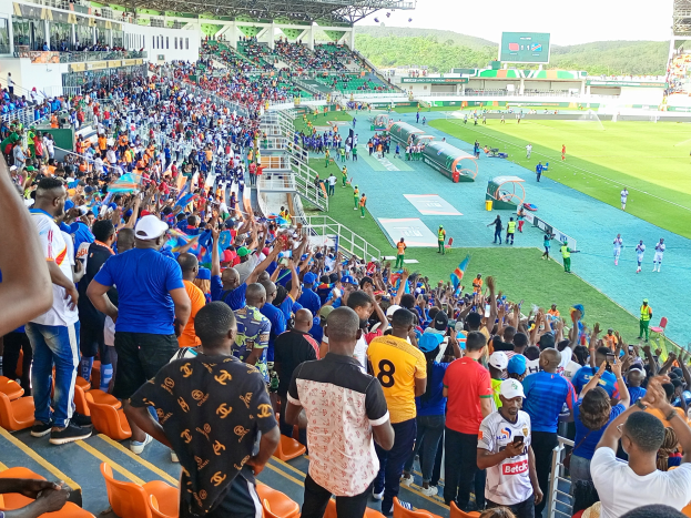 Eine große Menschenmenge versammelt sich vor einem eingezäunten Fußballfeld, wobei einige auf Stühlen sitzen und andere stehen, unter einem klaren blauen Himmel mit Hügeln im Hintergrund.