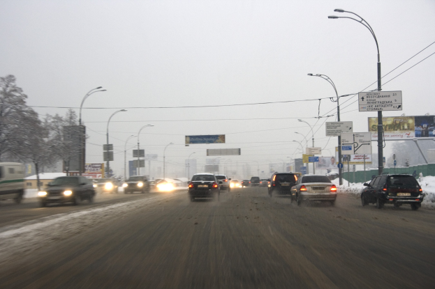 Eine belebte Stadtstraße an einem verschneiten Tag mit fahrenden Fahrzeugen, schneebedeckter Straße, Laternen, Texttafeln, Bäumen, Gebäuden und einem Himmel im Hintergrund.