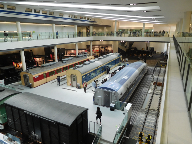 Eine geräumige Museumsausstellung zeigt verschiedene Züge in unterschiedlichen Größen und Farben, bei der Besucher vom Boden, Geländern und Säulen unter Deckenbeleuchtung zuschauen.