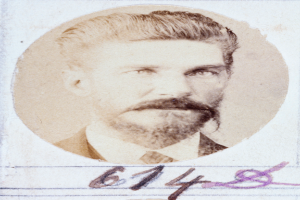 Ein altes Foto eines Mannes mit Bart und Schnurrbart, der direkt in die Kamera schaut und leicht geöffnete Lippen hat, ordentlich gekämmtes Haar und einen gut gepflegten Bart.