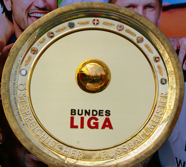 Ein Mann hält ein goldenes Schild mit "Bundesliga" darauf, mit einem Banner im Hintergrund mit drei Personen.