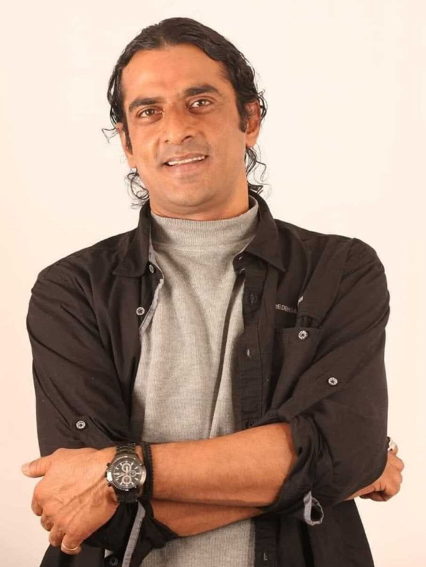 Ein Mann in einem schwarzen und aschfarbenen Kleid mit überschlagenen Armen, der vor einem weißen Hintergrund steht und eine Uhr tr├Ągt, der nachdenklich erscheint.